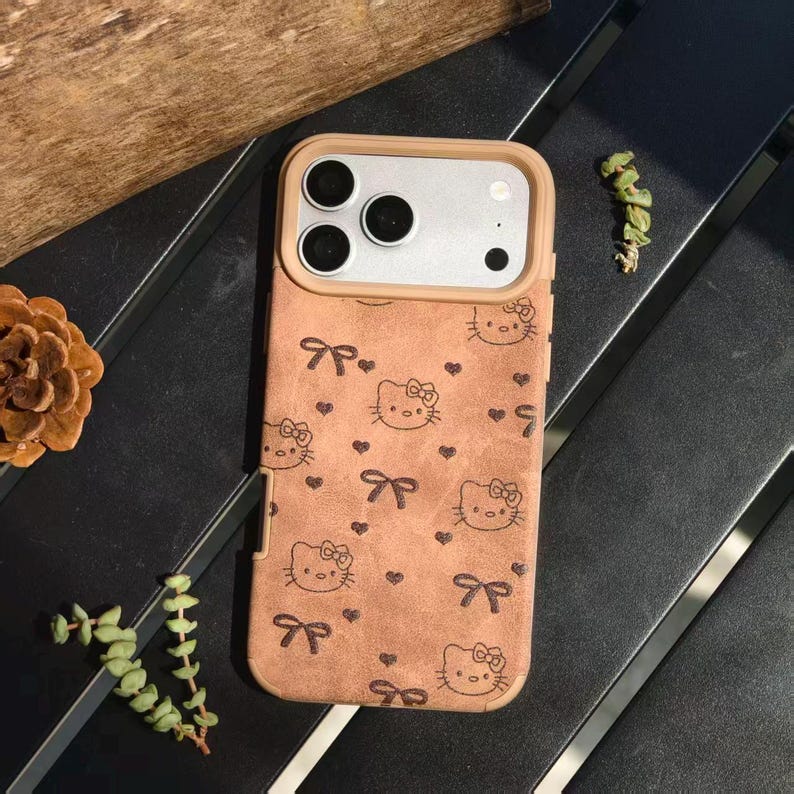 May include: A phone case with a tan suede-like texture, featuring a repeating pattern of Hello Kitty faces, bows, and hearts. The case has a light brown border around the camera and phone edges.