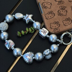 May include: A silver beaded bracelet with various charms, including a star with "PINK" text, a heart, and a cube with a bunny design. The bracelet features large, round beads and a rhinestone-studded bead. A brown phone case with a Hello Kitty pattern is in the background.