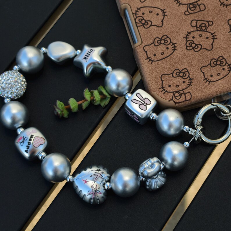 May include: A silver beaded bracelet with various charms, including a star with "PINK" text, a heart, and a cube with a bunny design. The bracelet features large, round beads and a rhinestone-studded bead. A brown phone case with a Hello Kitty pattern is in the background.