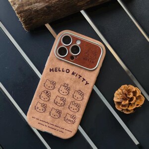 Op de afbeelding: Tan telefoonhoesje met Hello Kitty-ontwerpen. De hoes heeft een bruine, leerachtige textuur met meerdere Hello Kitty-gezichten en de woorden "HELLO KITTY". De camera is bruin met een metalen ring.