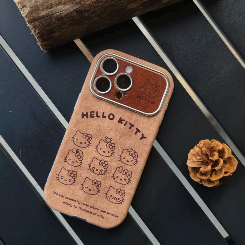 Op de afbeelding: Tan telefoonhoesje met Hello Kitty-ontwerpen. De hoes heeft een bruine, leerachtige textuur met meerdere Hello Kitty-gezichten en de woorden "HELLO KITTY". De camera is bruin met een metalen ring.