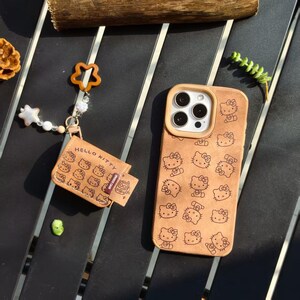 Peut inclure: Coque de t&eacute;l&eacute;phone et porte-cartes beiges orn&eacute;s de motifs Hello Kitty. L'&eacute;tui de t&eacute;l&eacute;phone a un bord marron clair autour de l'appareil photo. Le porte-cartes a une petite sangle avec un charme fleur et &eacute;toile. Le porte-cartes dit "Hello Kitty".