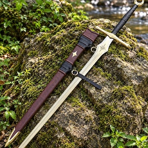 May include: A metal sword with a gold-toned crossguard and pommel, featuring a black leather-wrapped grip. The blade has a star-shaped engraving. It is accompanied by a brown leather scabbard with black leather straps and gold accents.