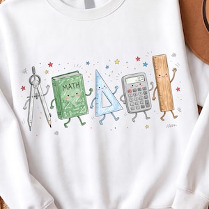 May include: A white crewneck sweatshirt featuring a colorful graphic of anthropomorphic school supplies including a compass, a green book labeled MATH, a blue triangle ruler, a grey calculator, and a wooden ruler, all surrounded by small stars.