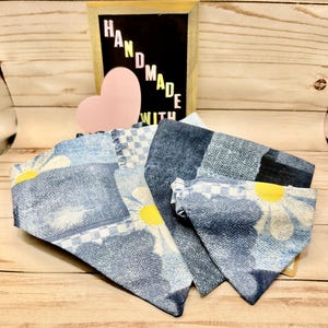 May include: Two triangular pet bandanas featuring a patchwork design of blue denim-style fabric, white and blue checkered patterns, and large yellow daisy motifs. A small pink heart and a sign with the text HANDMADE WITH are visible behind.
