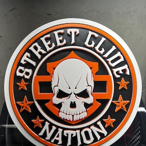 May include: A round, layered sign with the words "STREET GLIDE NATION" in white lettering. The sign features a skull graphic in the center, surrounded by orange stars and accents. The design is set against a black and orange background.