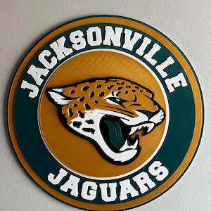 May include: A circular sign featuring the Jacksonville Jaguars logo. The design includes a jaguar head in black, white, and gold, set against a gold and teal background. The words "JACKSONVILLE JAGUARS" are in white lettering around the edge.