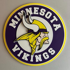 May include: A round Minnesota Vikings logo in purple, white, yellow, and black. The logo features a Viking head with a yellow helmet and beard, and the words "MINNESOTA VIKINGS" in white lettering around the edge.