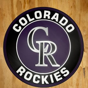May include: A circular sign featuring the Colorado Rockies logo with a purple center, a black border, and white text reading COLORADO ROCKIES. The central design displays a large, stylized silver and black CR monogram on a purple background.
