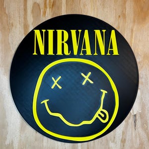 May include: A circular black wall sign featuring the yellow text NIRVANA at the top. Below the text is a yellow graphic of a smiley face with X-shaped eyes and a wavy mouth with a tongue sticking out, all set against a solid black background.