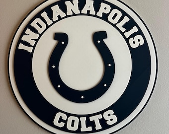 Könnte beinhalten: Ein kreisförmiges Indianapolis Colts-Logo in Marineblau, Weiß und Schwarz. Das Logo zeigt ein weißes Hufeisen in der Mitte, umgeben von den Wörtern "Indianapolis" und "Colts" in weißer, fetter Schrift. Das Logo ist auf einer hellgrauen Oberfläche montiert.