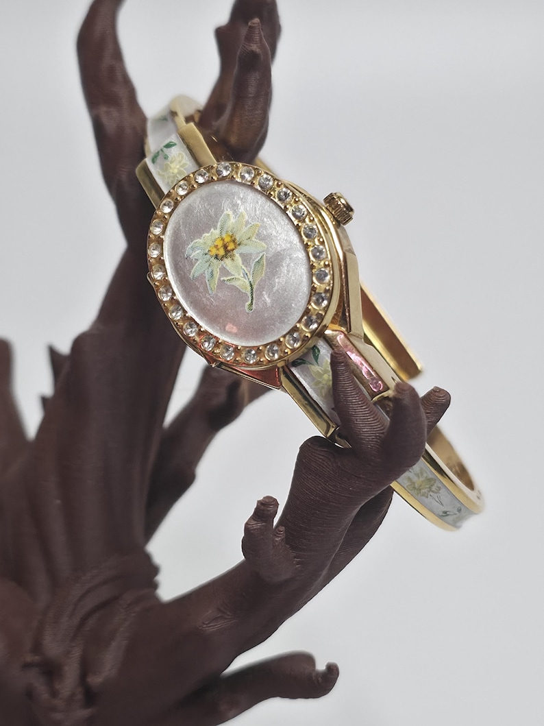 May include: Gold-coloured bangle watch with a white face featuring a floral design. The watch face is surrounded by a ring of clear crystals. The bangle has a floral pattern in white and yellow.