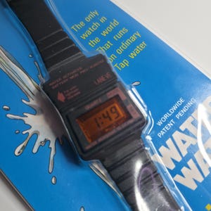 May include: A black digital watch with a black strap, displaying the time 1:49. The watch is packaged on a blue background with text that reads "The only watch in the world that runs on ordinary tap water."