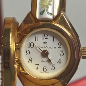 May include: Gold-plated Andr&eacute; Mouche quartz watch with a white face and black numerals. The watch has a floral design on the top and the words "Andr&eacute; Mouche" and "Swiss Made".