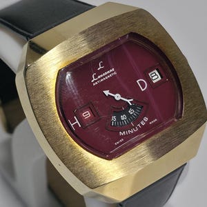 May include: A Lausanne Antimagnetic wristwatch with a gold-tone brushed metal case and a black leather strap. The watch features a deep red dial with a digital hour display showing 9, a minute arc, and a date window displaying 8.