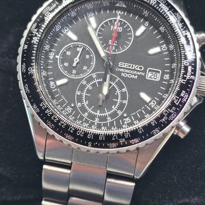 May include: A silver-tone stainless steel Seiko chronograph watch with a black dial, three sub-dials, a date window at the three o'clock position, and a rotating slide rule bezel featuring various conversion scales and markings.