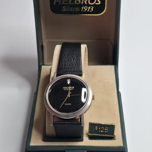 Könnte beinhalten: Eine Helbros Invincible Quarzuhr mit schwarzem Zifferblatt und Diamantakzent, präsentiert in einer grünen Box. Die Uhr hat ein schwarzes Lederarmband und ein silberfarbenes Gehäuse. Auf der Box steht "HELBROS Since 1913".