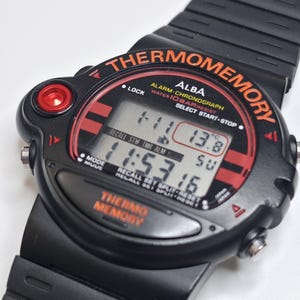 May include: Black Alba Thermomemory digital watch with a black band. The watch face has red and orange accents, with the text "THERMOMEMORY" and "ALBA". The digital display shows the time and temperature. The watch has multiple buttons and a red button on the side.