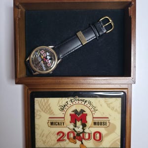 Może przedstawiać: Pamiątkowy zegarek na rękę Walt Disney World 2000 z czarnym skórzanym paskiem i złotą klamrą, umieszczony w drewnianym pudełku. Tarcza zegarka zawiera napis 2000 oraz postać Myszki Miki. Wieczko pudełka przedstawia napis Walt Disney World Mickey Mouse 2000.