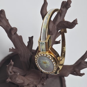 May include: A gold wristwatch featuring a floral design on the face and a band with white and green detailing. The watch face is surrounded by small, clear crystals. The watch is displayed on a brown, tree-like structure.