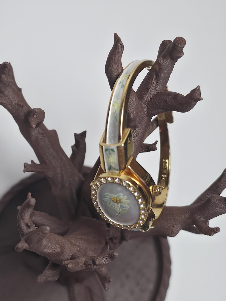 May include: A gold wristwatch featuring a floral design on the face and a band with white and green detailing. The watch face is surrounded by small, clear crystals. The watch is displayed on a brown, tree-like structure.