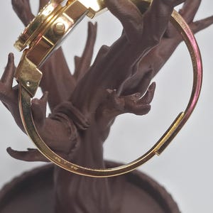 May include: A gold-coloured bangle watch with a round face and a bezel set with small, clear crystals. The watch is displayed on a brown, tree-like decorative stand. The watch band is a hinged, open design.