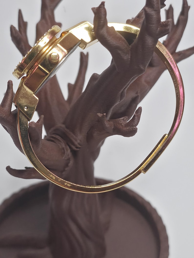 May include: A gold-coloured bangle watch with a round face and a bezel set with small, clear crystals. The watch is displayed on a brown, tree-like decorative stand. The watch band is a hinged, open design.