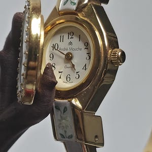 May include: Gold-coloured Andr&eacute; Mouche quartz watch with a white face and floral detailing. The watch features a hinged cover and a brown strap. The watch face shows the time with black numerals and the words "Andr&eacute; Mouche" and "Swiss Made".