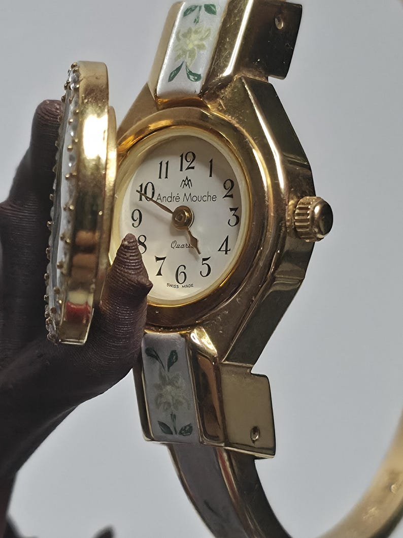 May include: Gold-coloured Andr&eacute; Mouche quartz watch with a white face and floral detailing. The watch features a hinged cover and a brown strap. The watch face shows the time with black numerals and the words "Andr&eacute; Mouche" and "Swiss Made".