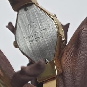 May include: A gold-toned Andr&eacute; Mouche Swiss Made watch. The watch has a rectangular face with a silver-coloured back. The watch band is also gold-toned. The watch is shown against a brown background.