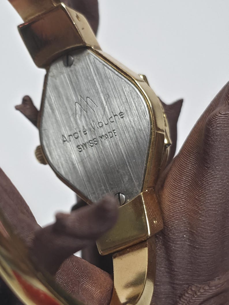 May include: A gold-toned Andr&eacute; Mouche Swiss Made watch. The watch has a rectangular face with a silver-coloured back. The watch band is also gold-toned. The watch is shown against a brown background.