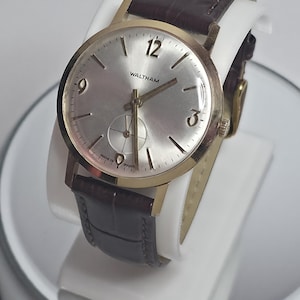 Op de afbeelding: Een goudkleurig Waltham polshorloge met een zilveren wijzerplaat en een bruine leren band. De wijzerplaat toont de cijfers 12, 3 en 9. Het horloge wordt gepresenteerd op een witte standaard. De woorden "MADE IN FRANCE" zijn zichtbaar op de wijzerplaat.