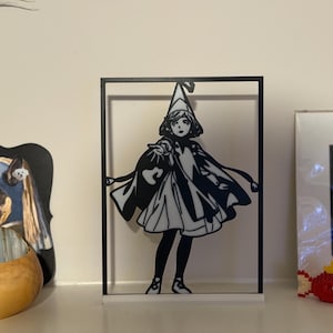 May include: Black and white silhouette of a figure in a framed display. The figure is wearing a hat and cape, holding a bag. The display is on a white shelf with other decorative items.