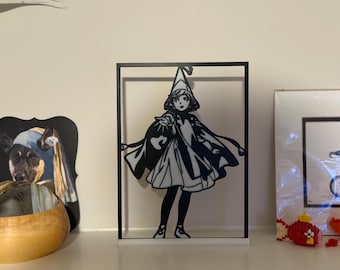 May include: Black and white silhouette of a figure in a framed display. The figure is wearing a hat and cape, holding a bag. The display is on a white shelf with other decorative items.