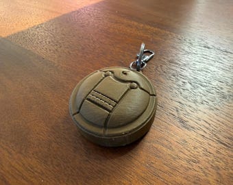 May include: A brown, circular keychain with a detailed design. The keychain features a textured surface with a central rectangular element and a metal clasp for attachment. The design is 3D printed.