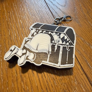 May include: A keychain featuring a cartoon illustration of a person reaching into a treasure chest. The chest is black and white with a brown interior. The person is white with black shoes.