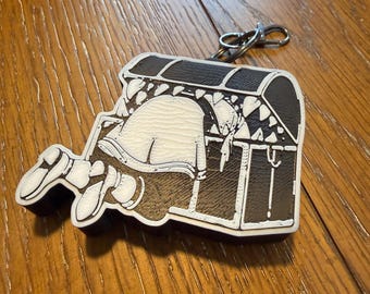 May include: A keychain featuring a cartoon illustration of a person reaching into a treasure chest. The chest is black and white with a brown interior. The person is white with black shoes.
