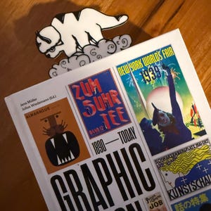 May include: A white enamel pin of a bison-like creature with black markings resting on clouds, positioned above a hardcover book titled Graphic Design 1890-Today, which features various vintage poster illustrations on its white cover.