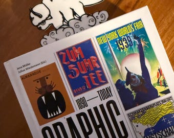 May include: A white enamel pin of a bison-like creature with black markings resting on clouds, positioned above a hardcover book titled Graphic Design 1890-Today, which features various vintage poster illustrations on its white cover.