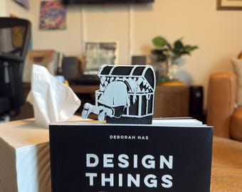 May include: A black and white bookmark featuring a person looking into a treasure chest. The bookmark is placed on top of a book with the title "DESIGN THINGS" and the author's name, DEBORAH NAS, in white text.