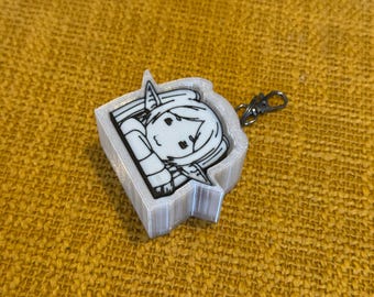 May include: A white and black cartoon character keychain with a silver-colored clasp. The keychain has a 3D-printed white border. The cartoon character has pointed ears and is wearing a hat. The keychain is on a yellow textured surface.