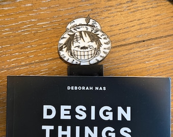 May include: A bookmark featuring a smiling cartoon character with a straw hat, placed on top of a book. The book has a black cover with the title "DESIGN THINGS" and the author's name, "DEBORAH NAS."