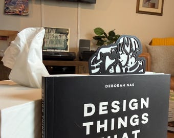 May include: A black and white anime-style bookmark featuring a character design. The bookmark is placed on top of a black book with the title "DESIGN THINGS THAT" in large white letters. The book's author is Deborah Nas.