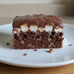 May include: A square slice of chocolate dessert on a white plate. The dessert has a dark brown, cake-like base with visible nuts, topped with a layer of white marshmallows and a dark chocolate frosting. A few crumbs and a marshmallow are on the plate.