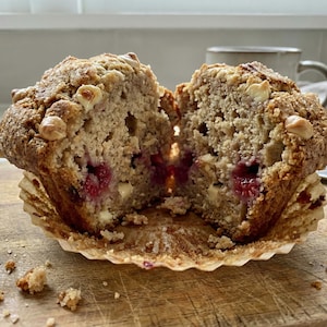 May include: A halved muffin, showing a cross-section of the baked treat with raspberries and white chocolate. The muffin sits in its paper case on a wooden board, with crumbs visible. A cup is in the background.