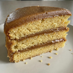 Puede incluir: Una porción de pastel en capas con un bizcocho amarillo dorado y un relleno y glaseado color caramelo. El pastel está sobre un plato blanco, con migas visibles. Un postre delicioso.
