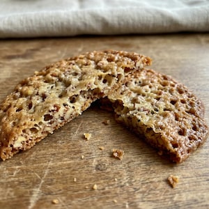 Könnte beinhalten: Zwei Hälften eines zerbrochenen, dünnen, knusprigen Kekses mit goldbrauner Farbe und strukturierter Oberfläche. Der Keks liegt auf einer Holzoberfläche, mit einem neutralfarbenen Tuch im Hintergrund. Der Keks scheint eine Art Krokant oder Spitzkuchen zu sein.