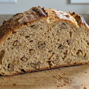 Può includere: Una pagnotta di pane alle noci appena sfornata, tagliata per rivelare l'interno. Il pane ha una crosta dorata e marrone ed è cosparso di pezzi di noci visibili. È appoggiato su un tagliere di legno, con briciole sparse.