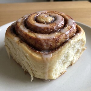 Könnte beinhalten: Ein frisch gebackener Zimtschnecken mit Zimtwirbel und einem großzügigen Guss aus weißem Zuckerguss. Das Gebäck liegt auf einem weißen Teller und zeigt sein goldbraunes Äußeres und seine weichen, fluffigen Schichten. Ein köstlicher Genuss.