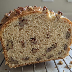 Könnte beinhalten: Ein teilweise geschnittenes Brot mit Pekannüssen, das auf einem Metall-Kühlrost liegt. Das Brot hat eine hellbraune Kruste und ein weiches, poröses Inneres. Pekannüsse sind oben und im Brot sichtbar. Das Brot sieht frisch gebacken aus.